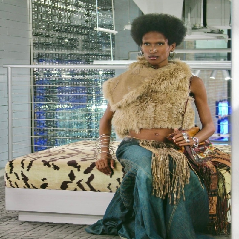 Black non-binary queer wearing long deim skirt, fur vest and fringe tassel beads. 