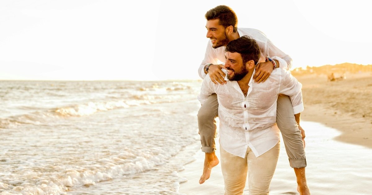 Gay male couple walking piggy back style on the beach