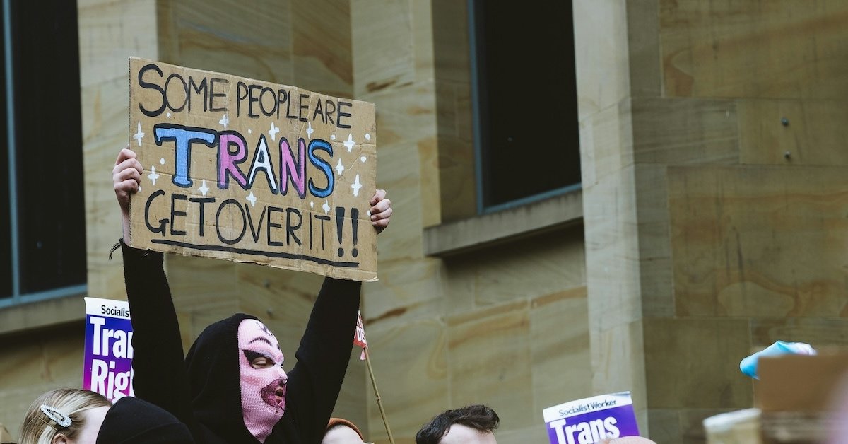 A person holding a sign that reads "Some People are Trans, Get Over It."