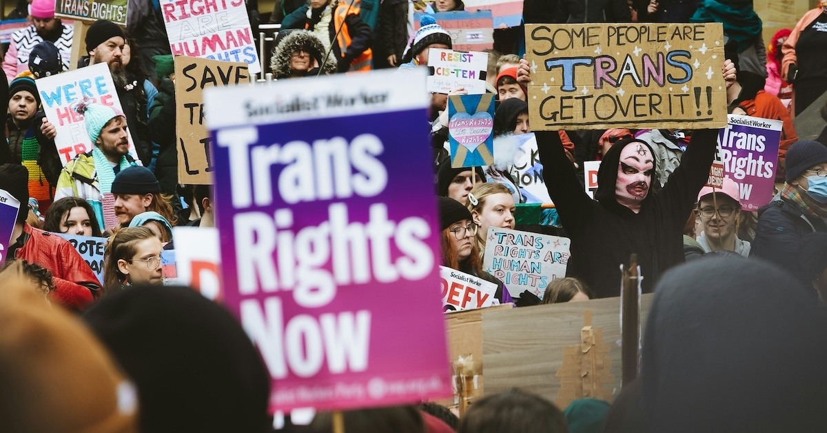 Protestors with signs saying "SOME PEOPLE ARE TRANS GET OVER IT" and "Trans Rights Now."