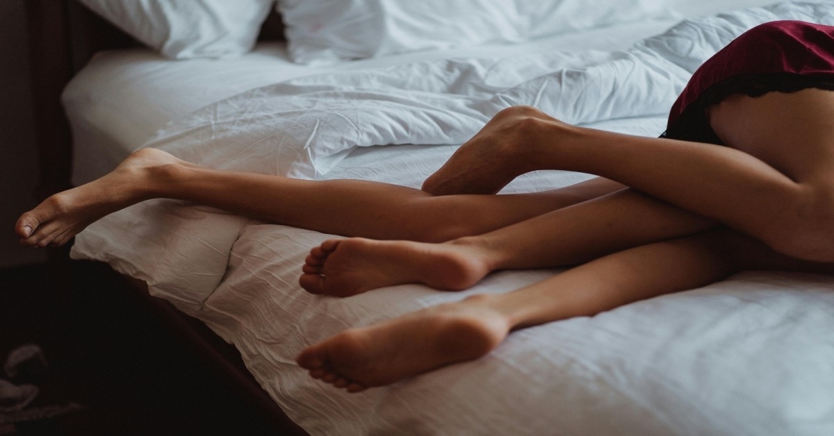 Two people from the hips and knees down with bare feet entangled while lying on a white bed.
