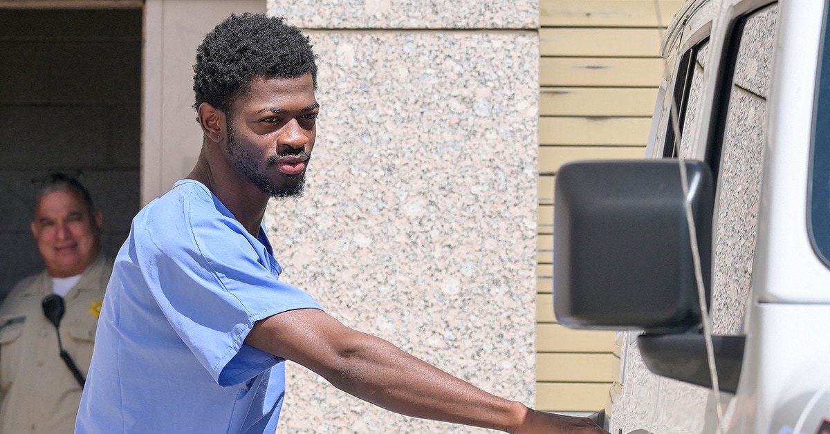Lil Nas X reaching for a van door handle outside of a prison; a police officer stands behind him